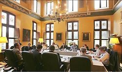 In de grote vergaderzaal zitten de onderhandelaars aan tafels die in een vierkant staan. De vier partijen hebben elk drie onderhandelaars. Photo News<br>