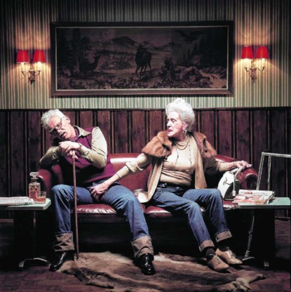 Een oudere vrouw, haren netjes in een chignon, beige twinset, maar met een strakke jeans aan, grijpt haar slapende man in het kruis van zijn jeans. Roldoorbrekend of spottend?Erwin Olaf